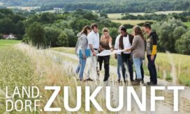 Acht junge Menschen stehen auf einem gepflasterten Feldweg in einer Wald- und Wiesenlandschaft und betrachten einen Plan, den sie in den Händen halten.